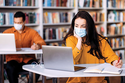 Photo of College students stitting far apart looking at laptops with face masks on.