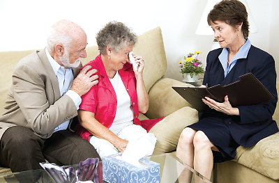 Photo: An elderly couple is seated on a couch, speaking with a therapist who is taking notes. The elderly woman is crying. 