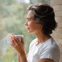 Photo: lady close to a window with a cup in her hands looking outside