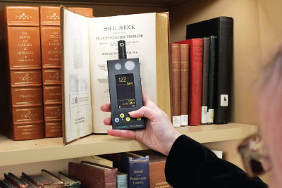 Bexx Caswell-Olson, M.S.L.I.S., holds up a light meter to a shelf of antique books in the Melvin Sabshin, M.D. Library & Archives to measure the amount of visible and infrared light to which the books are exposed.