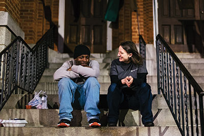 Photo of Liz Frye, M.D., M.P.H., and an homeless man sitting on a stoop in Pittsburgh.
