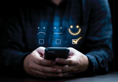 Photo of person using a phone with three face emojis floating above. one sad, one expressionless, and one smiling. The smiling face is glowing and has a check mark below it.
