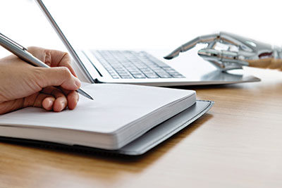 Image of a human hand taking notes with a robot hand using a laptop.