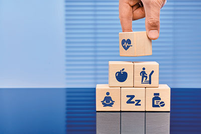 Photo of wooden blocks stacked in a pyramid with various integrated health icons printed on them and a hand placing the last block on top.